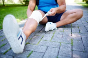 Close-up of an athlete sitting on a paved path, carefully wrapping his knee with a white bandage, highlighting the importance of proper care during injury recovery.