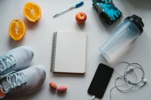 A flat lay of essential training resources including a notebook, pen, sneakers, a water bottle, fruit, nuts, and workout gloves, emphasizing the importance of planning and proper nutrition in fitness.
